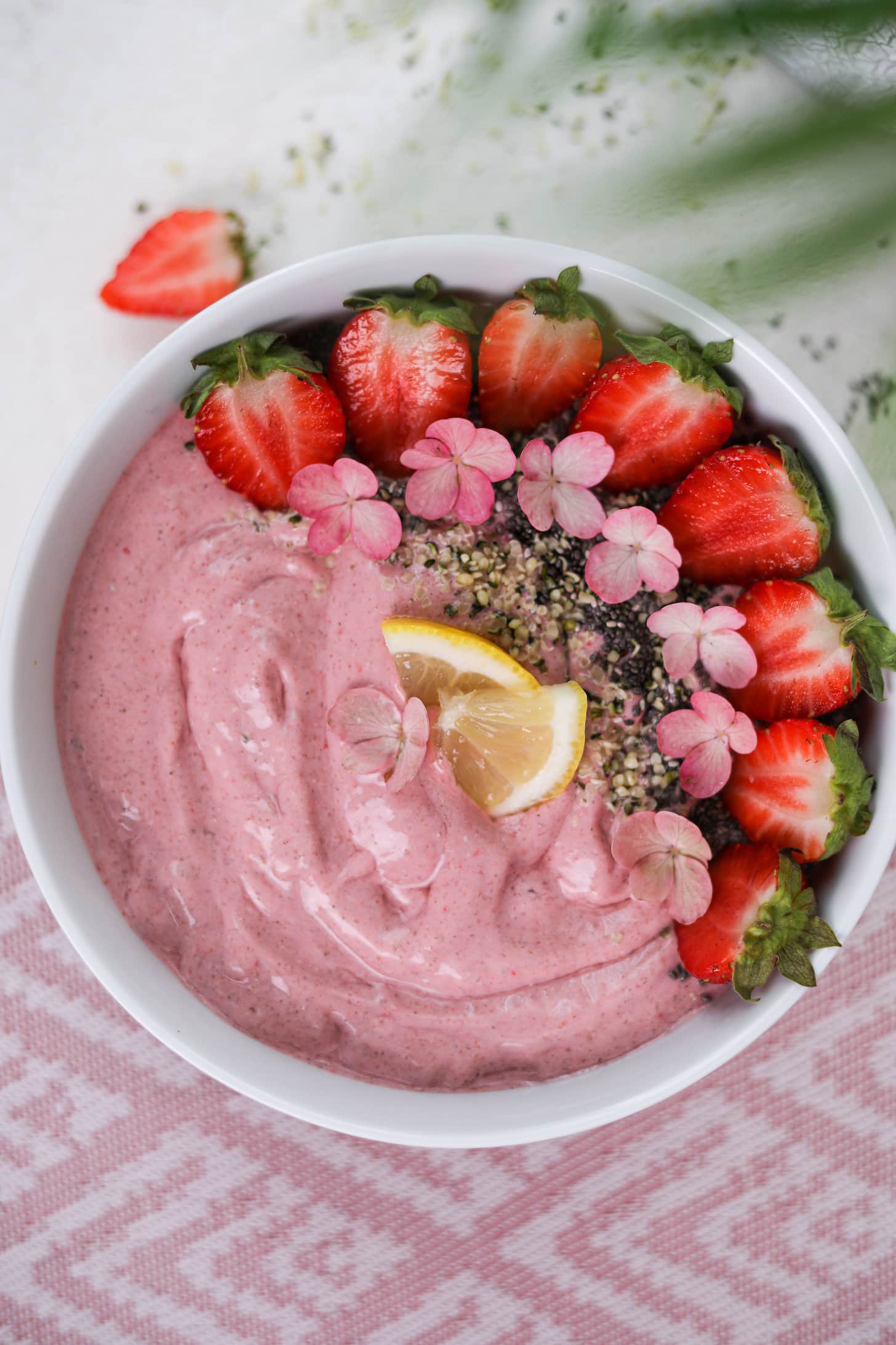 Overhead of a pink smoothie topped with strawberry halves, pink flowers and seeds.