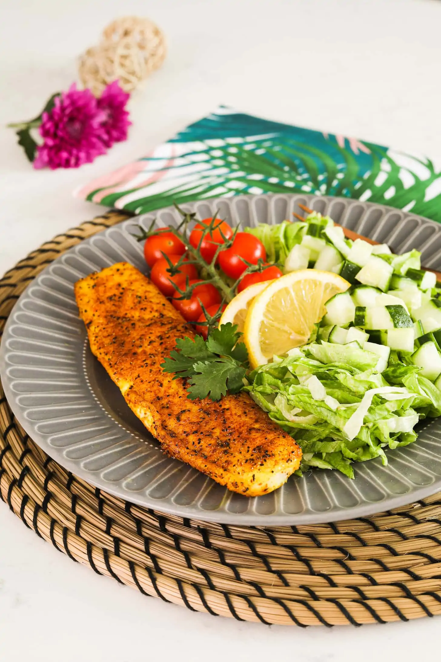 A plate of fish fillet with a side of chopped lettuce, cucumber slices, baby tomatoes on a vine and lemon slices.