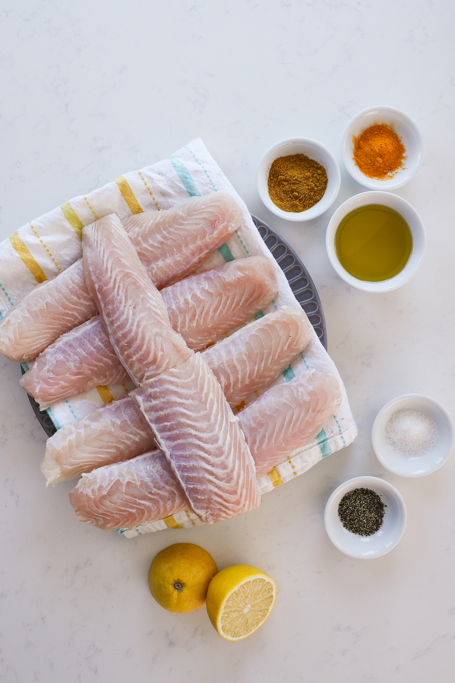 A collection of ingredients including white fish fillets, spices, oil and lemon halves.