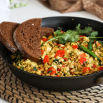 A fry pan of scramble with red pepper cubes topped with cilantro and green chilli and two slices of pumpernickel bread placed on top.