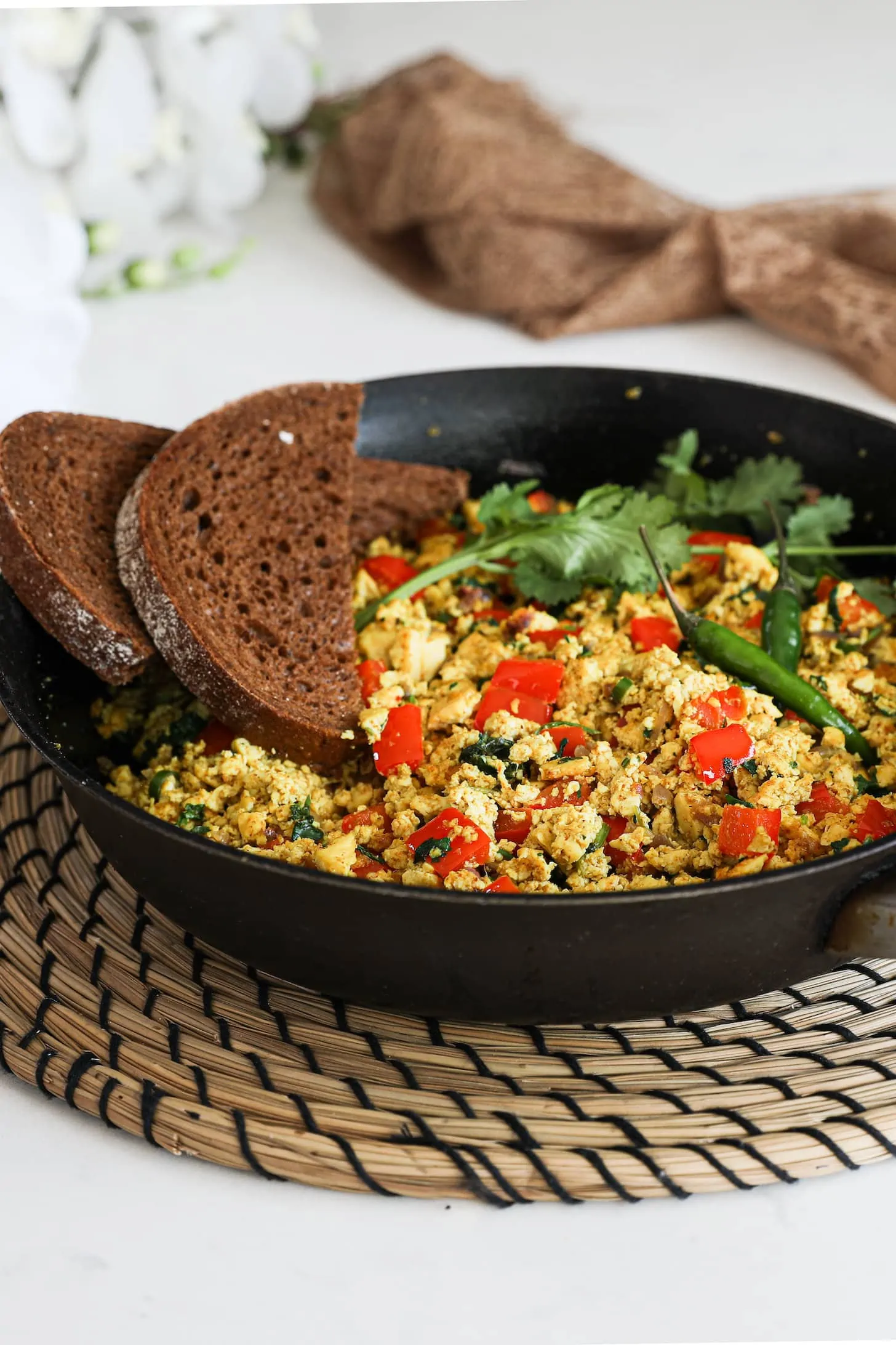 A fry pan of scramble with red pepper cubes topped with cilantro and green chilli and two slices of pumpernickel bread placed on top.