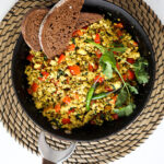 Overhead shot of a pan of scramble with red pepper cubes topped with cilantro and green chilli and two slices of pumpernickel bread placed on top.