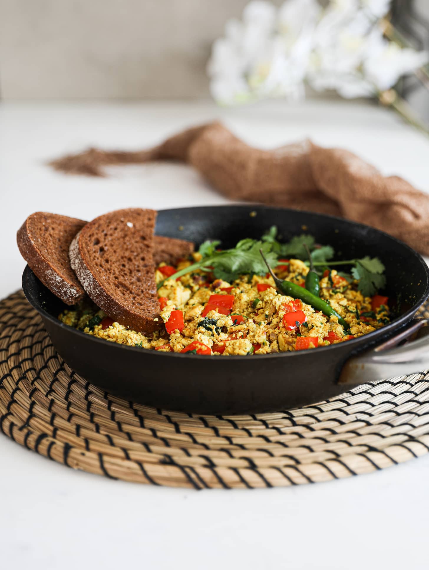 Perspective image of a fry pan of scramble with red pepper cubes topped with cilantro and green chilli and two slices of pumpernickel bread placed on top. There are flowers in the background.