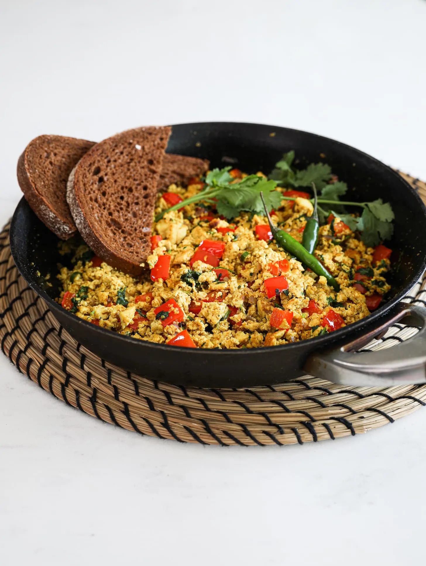 Perspective image of a fry pan of scramble with red pepper cubes topped with cilantro and green chilli and two slices of pumpernickel bread placed on top. The pan is placed on a rope place mat.