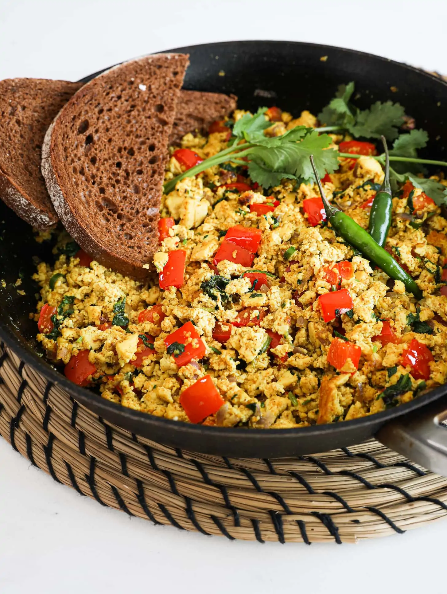 Perspective image of a fry pan of scramble with red pepper cubes topped with cilantro and green chilli and two slices of pumpernickel bread placed on top. Pan is placed on a rope place mat.