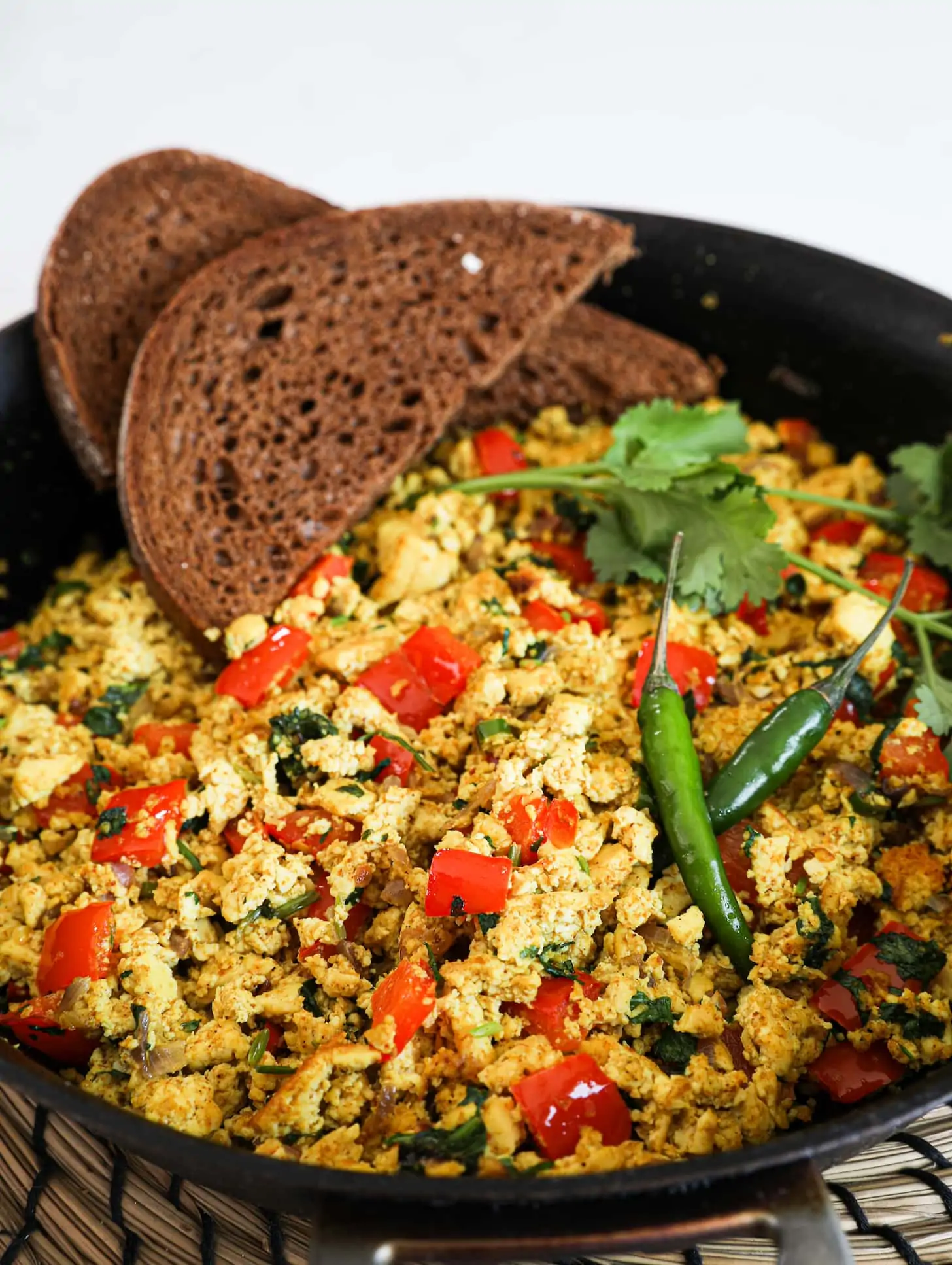 A close up image of a fry pan of scramble with red pepper cubes topped with cilantro and green chilli and two slices of pumpernickel bread placed on top.