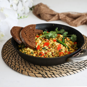A pan of scramble with red pepper cubes topped with cilantro and green chilli and two slices of pumpernickel bread placed on top. There is a brown scarf and flowers in the background.