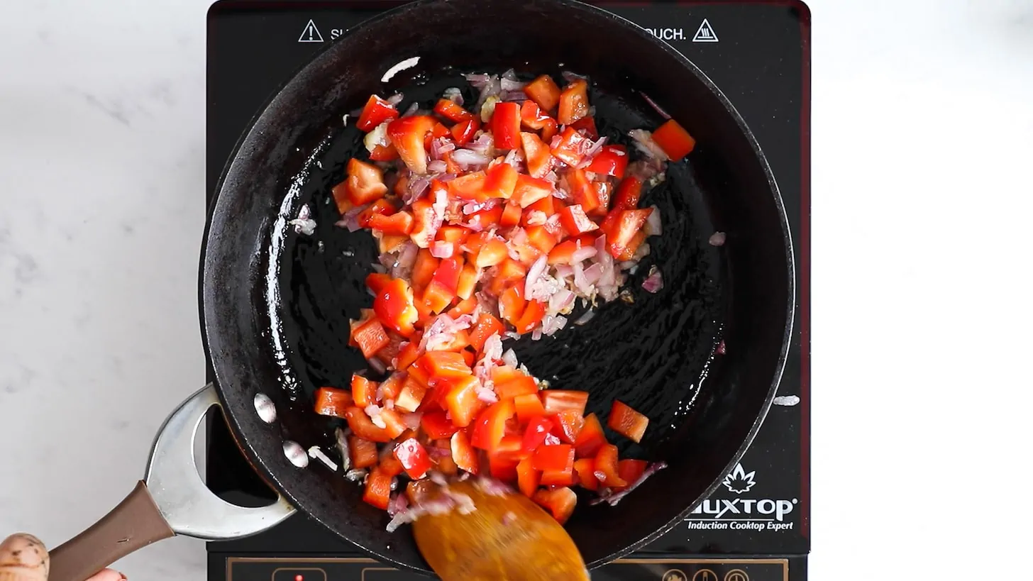 A hand holding a wooden spoon and stirring cubes of red pepper and chopped onion in a pan.