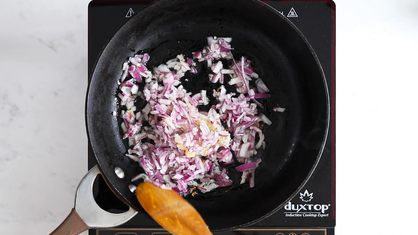 A hand holding a wooden spoon and stirring chopped onion in a pan.