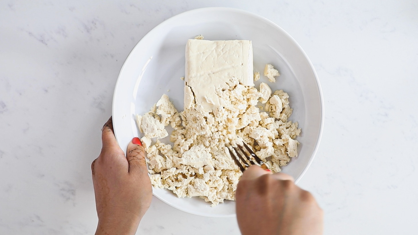A hand holding a fork mashing a block of tofu in a bowl.