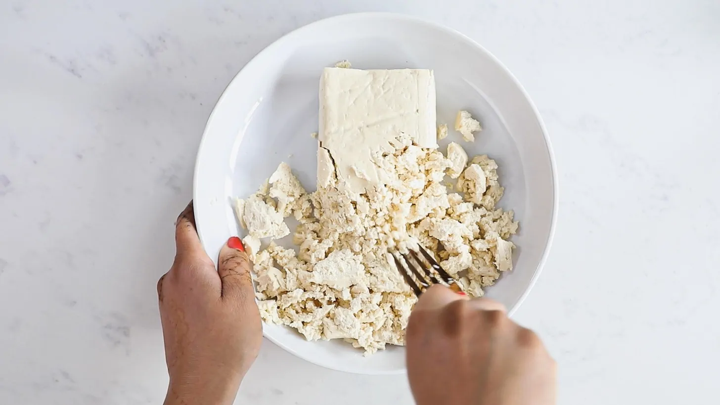 A hand holding a fork mashing a block of tofu in a bowl.
