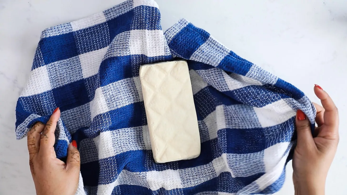Two hands wrapping a block of tofu in a blue checkered t-towel.