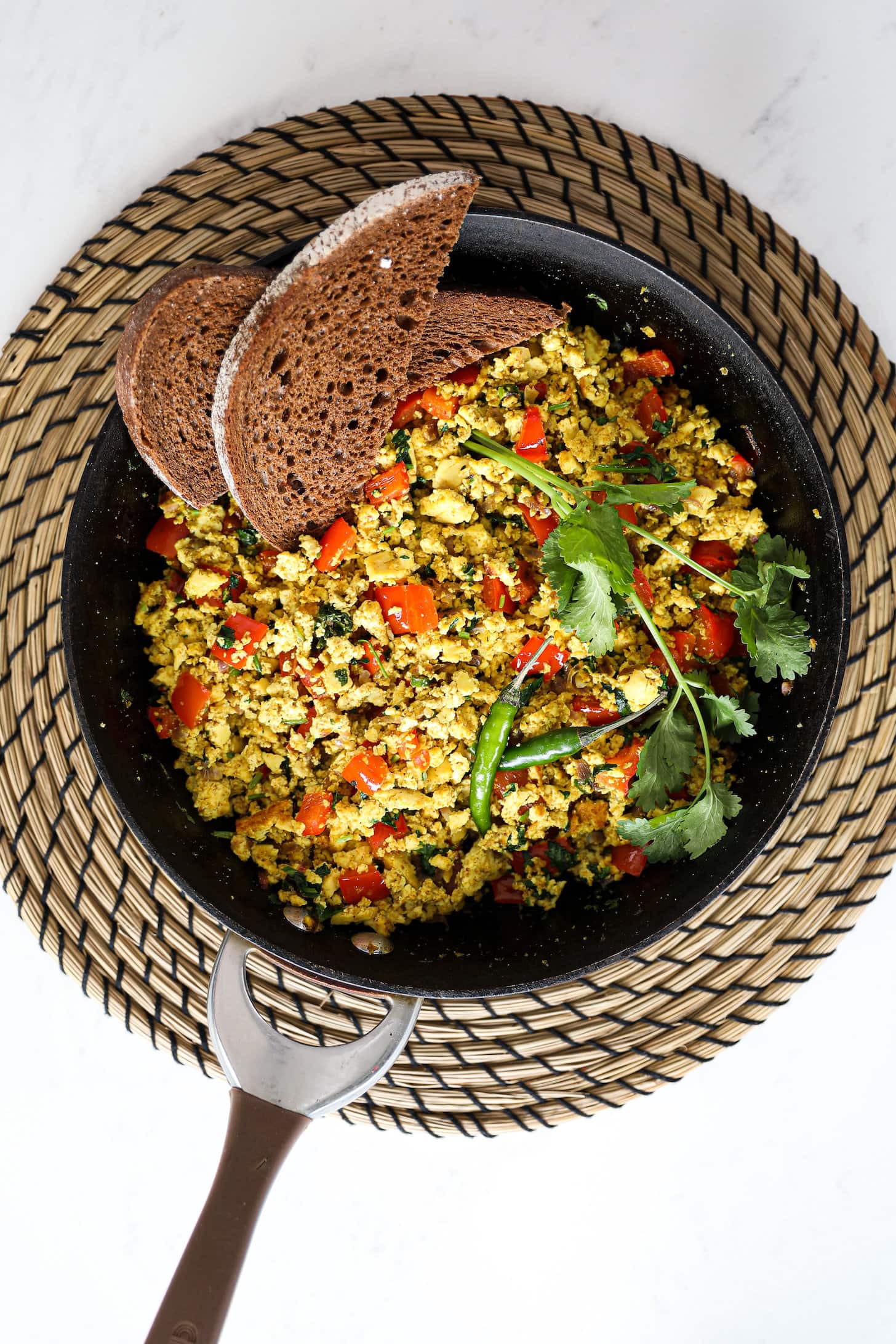 Overhead shot of a pan of scramble with red pepper cubes topped with cilantro and green chilli and two slices of pumpernickel bread placed on top.