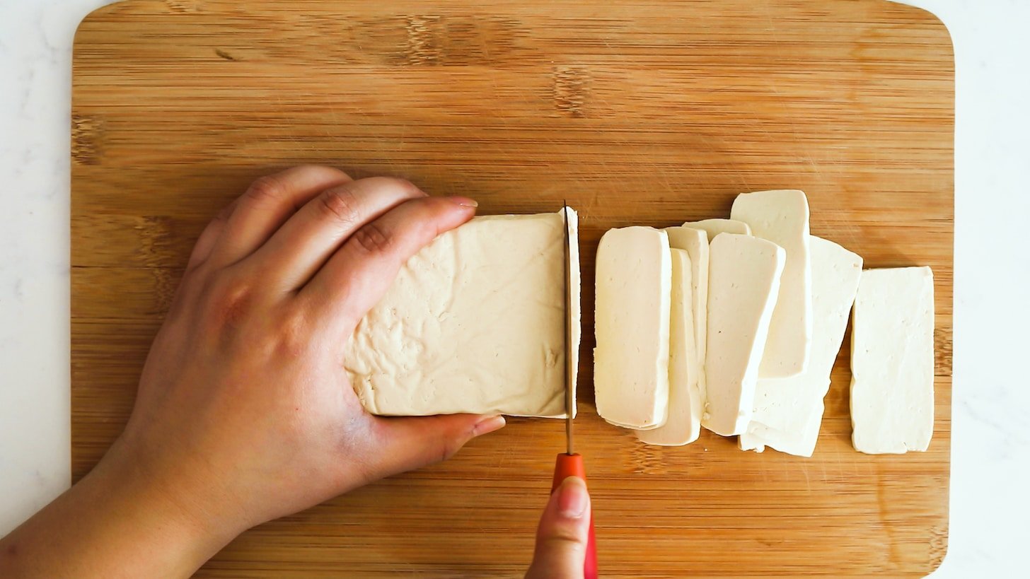 A hand holding a block of tofu whilst slicing it in slabs with the other hand.