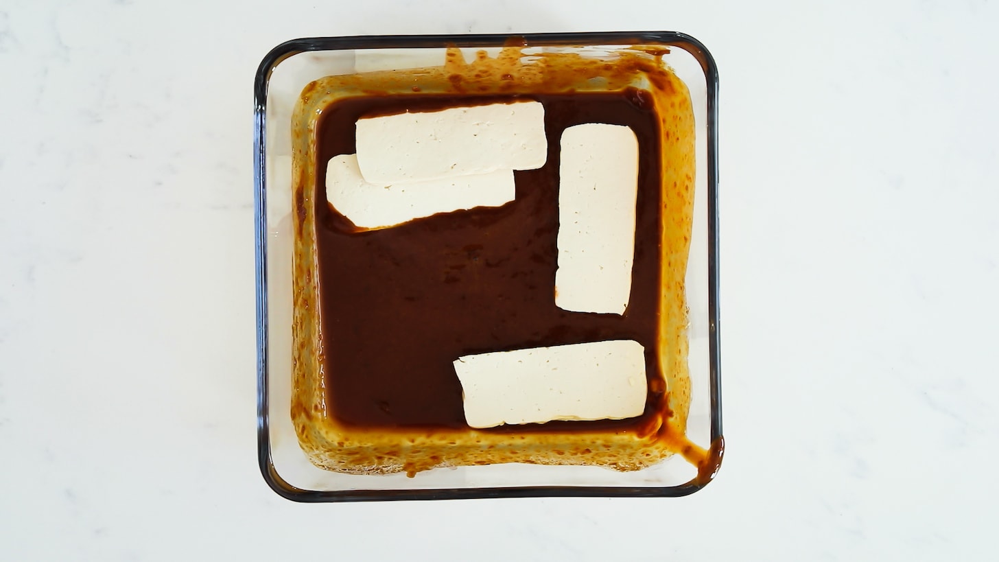 A container with a brown sauce with a handful of tofu slabs in it.