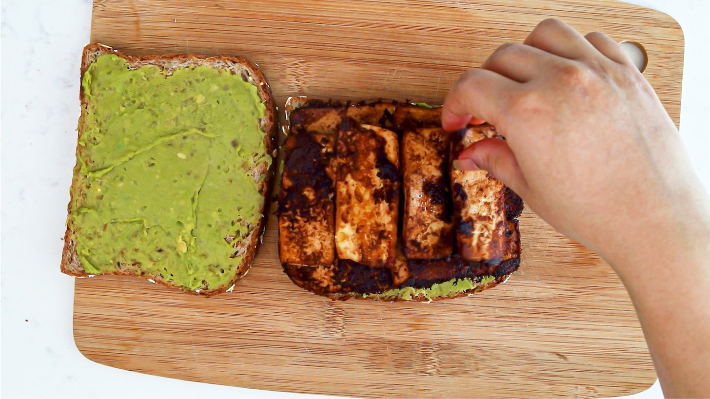 A hand arranging slabs of tofu on a slice of bread coated in a green spread.