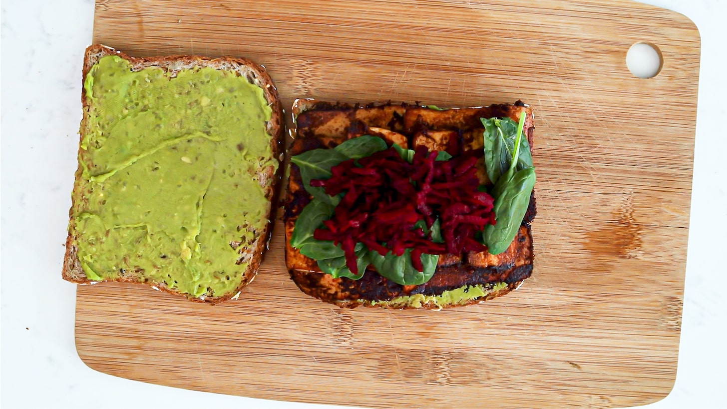 Two slices of bread, and one is topped with slabs of tofu, spinach and grated beetroot.