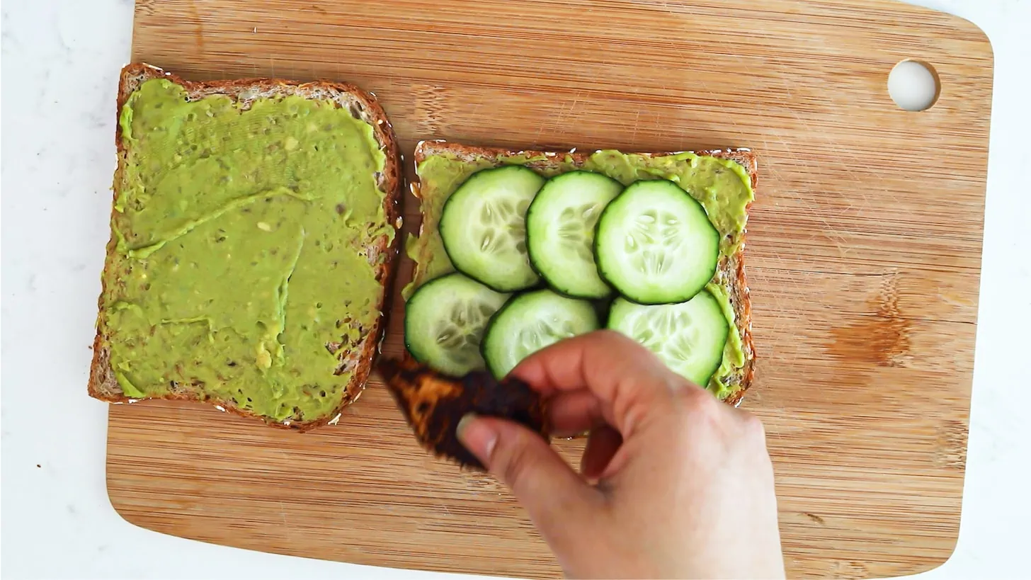 Two slices of bread with a green spread with one slice topped with cucumber slices.