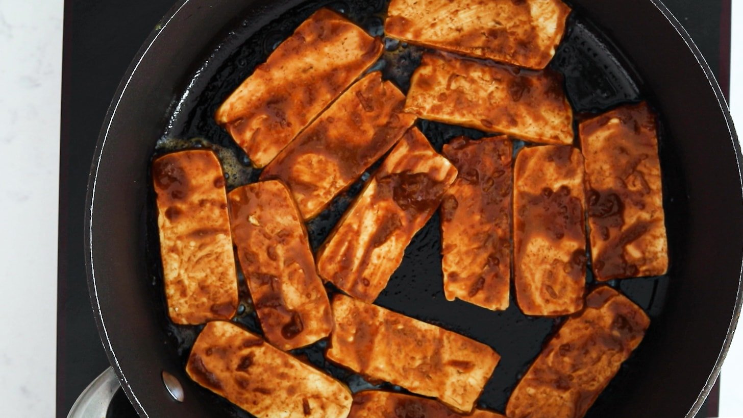 Slabs of tofu coated in a brown sauce cooking in a pan.