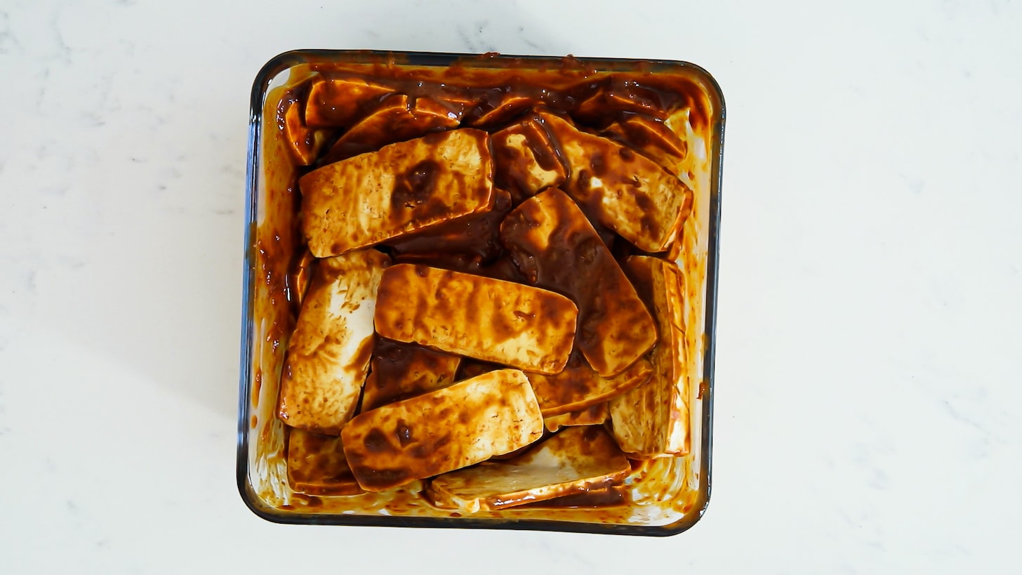 Slabs of tofu coated in a thick brown marinade in a glass container.