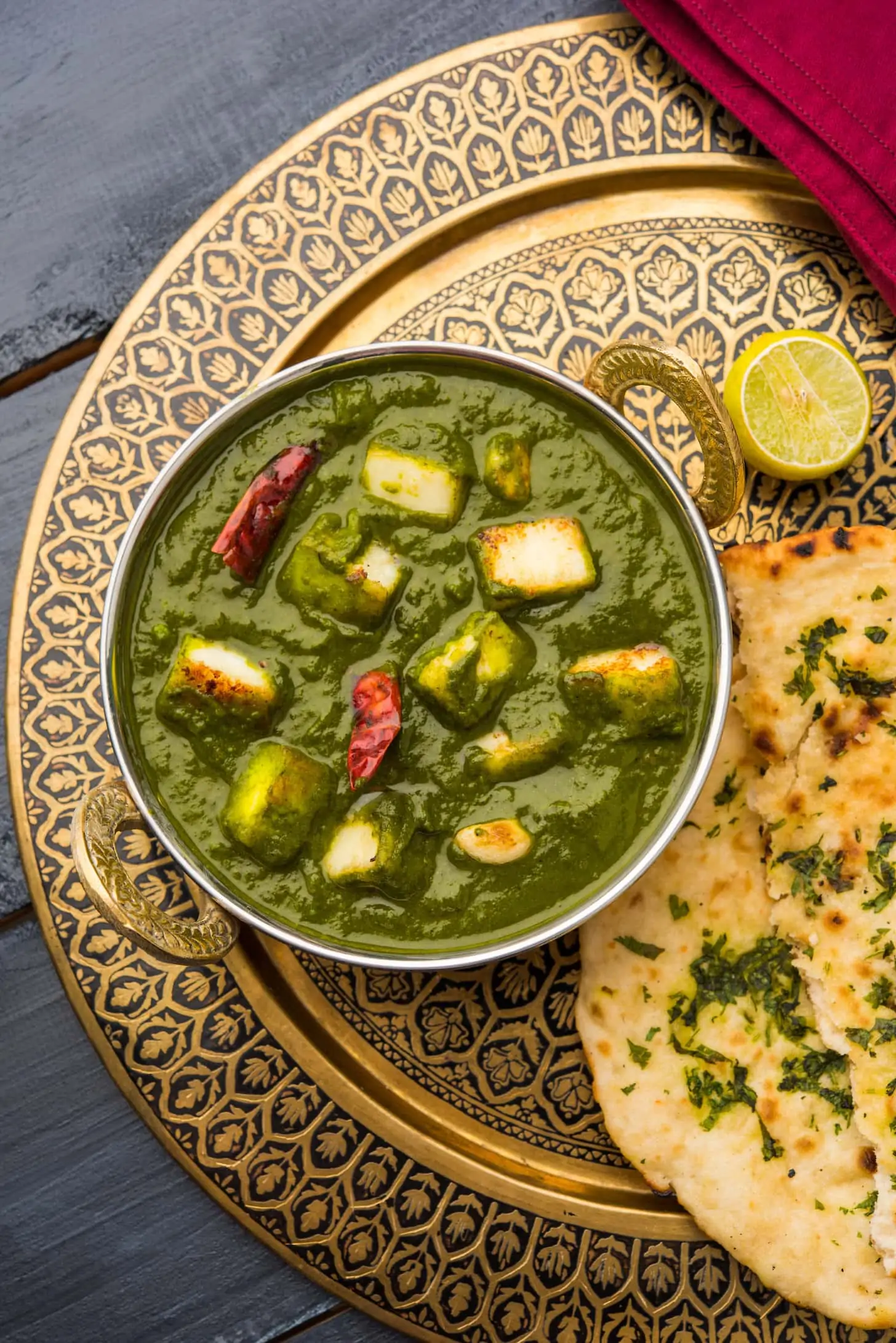 a bowl of green sauce with pieces of paneer on a patterned gold tray with a naan.
