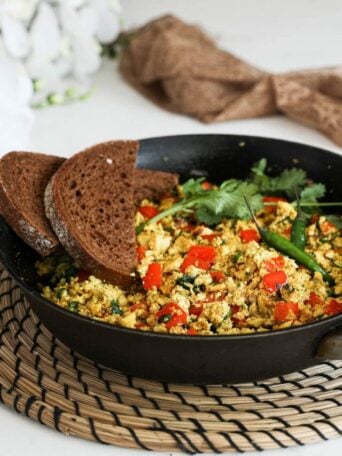 A fry pan of scramble with red pepper cubes topped with cilantro and green chilli and two slices of pumpernickel bread placed on top - perspective image.