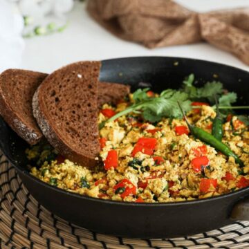 A fry pan of scramble with red pepper cubes topped with cilantro and green chilli and two slices of pumpernickel bread placed on top - perspective image.