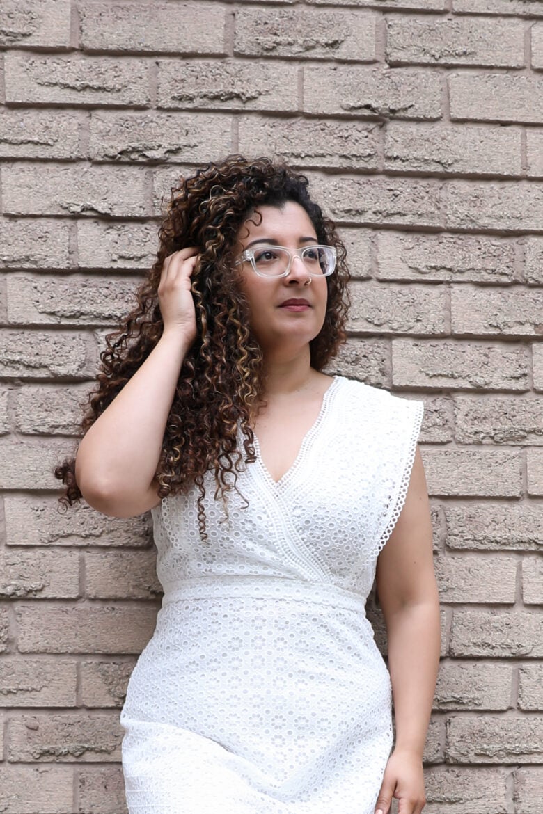 A curly-haired woman touching her hair leaning against a wall in a white dress.