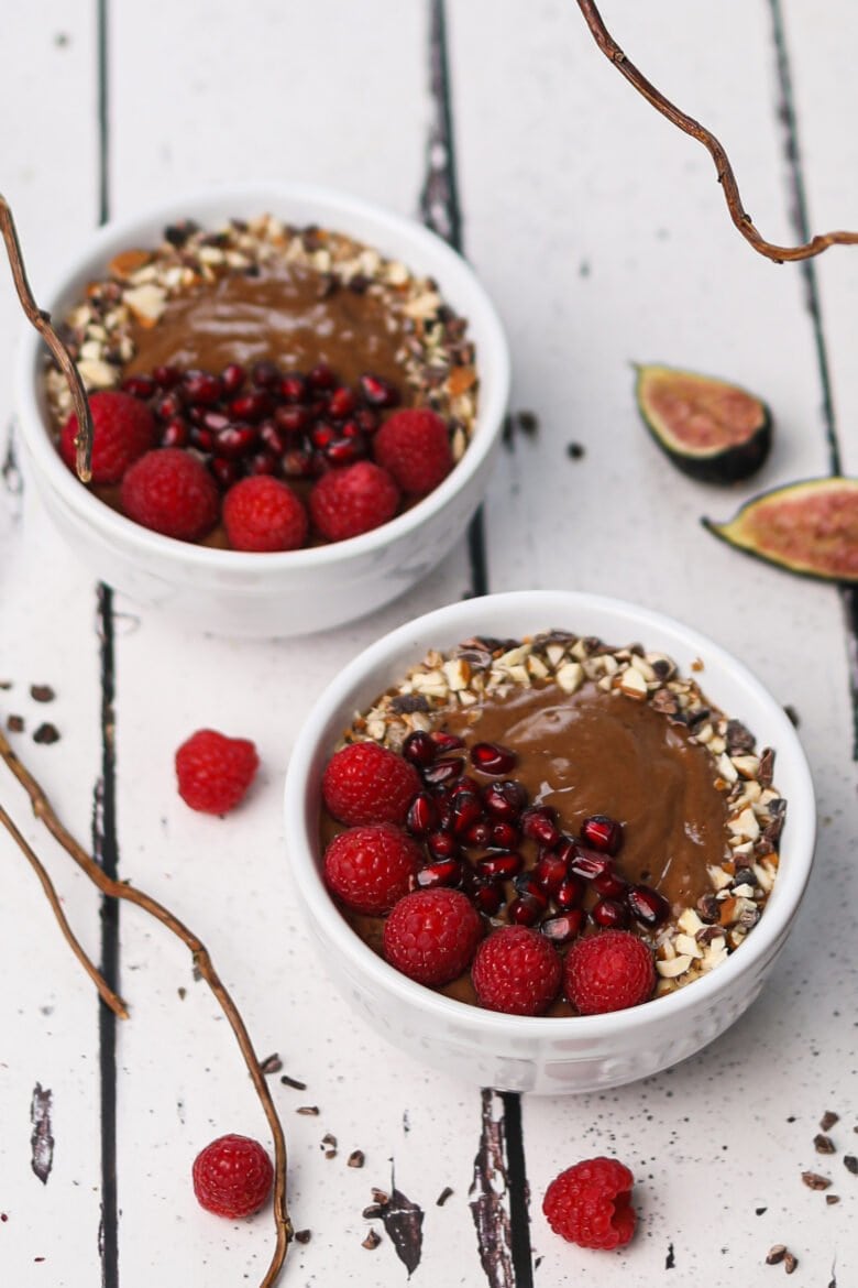 Perspective image of 2 bowls of chocolate smoothie topped with berries, nuts and pomegranate.