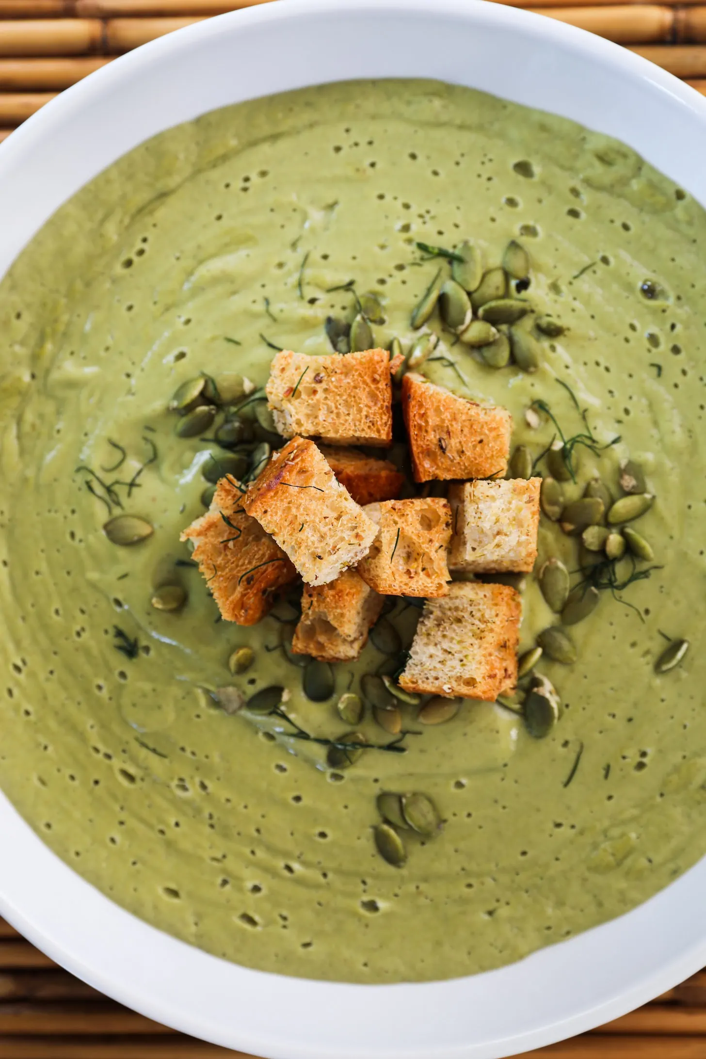 Flat lay of a bowl of green soup topped with a heap of croutons and seeds.