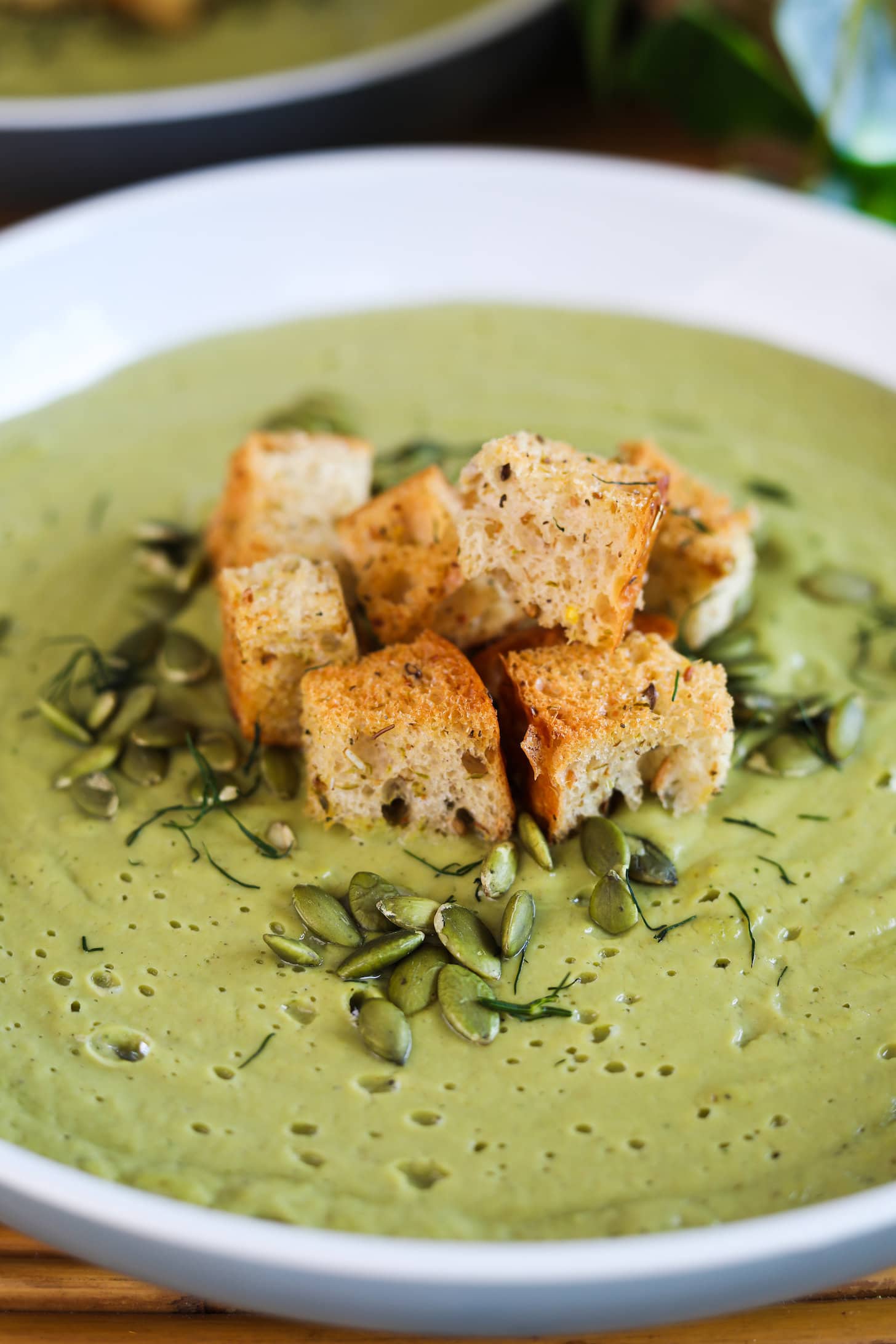 Angled perspective image of a bowl of green soup topped with croutons, seeds and herbs.