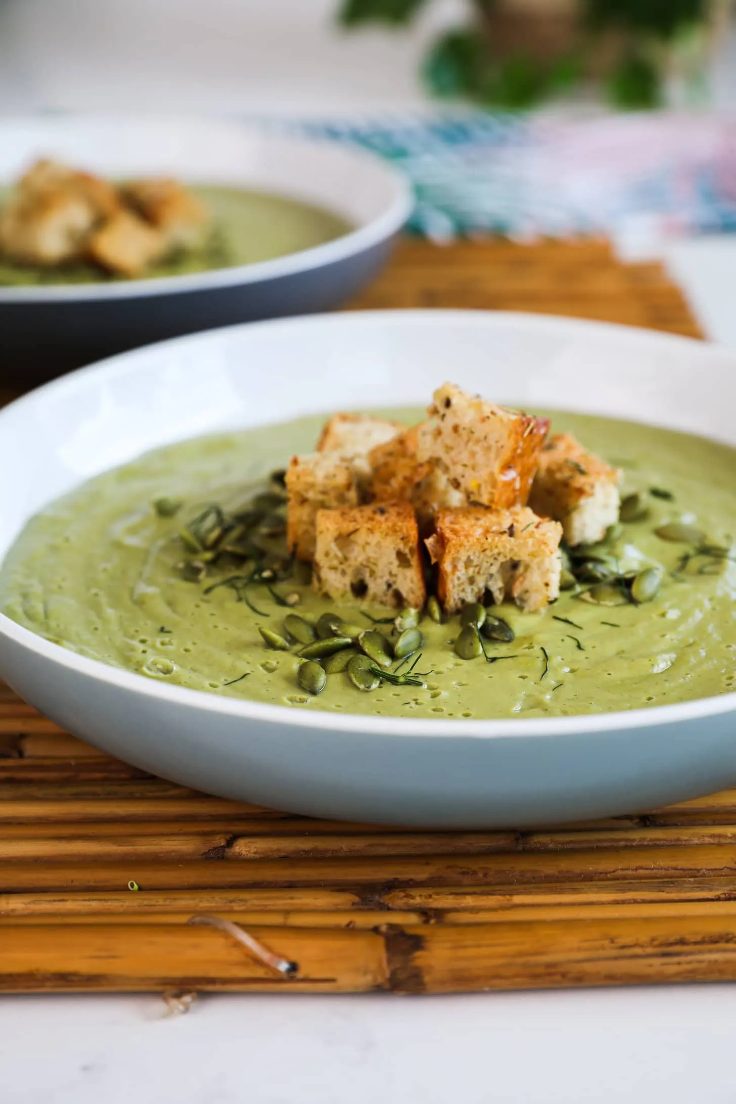 Perspective image of a bowl of green soup topped with croutons and seeds.