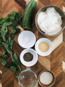 An array of food ingredients including half a cucumber, bowl of yogurt, and ramekins of spices and herbs on a wooden board.