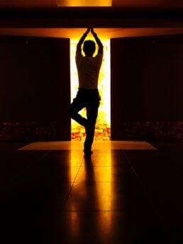 A person doing a yoga tree pose in the dark with a yellow light source entering through a door.