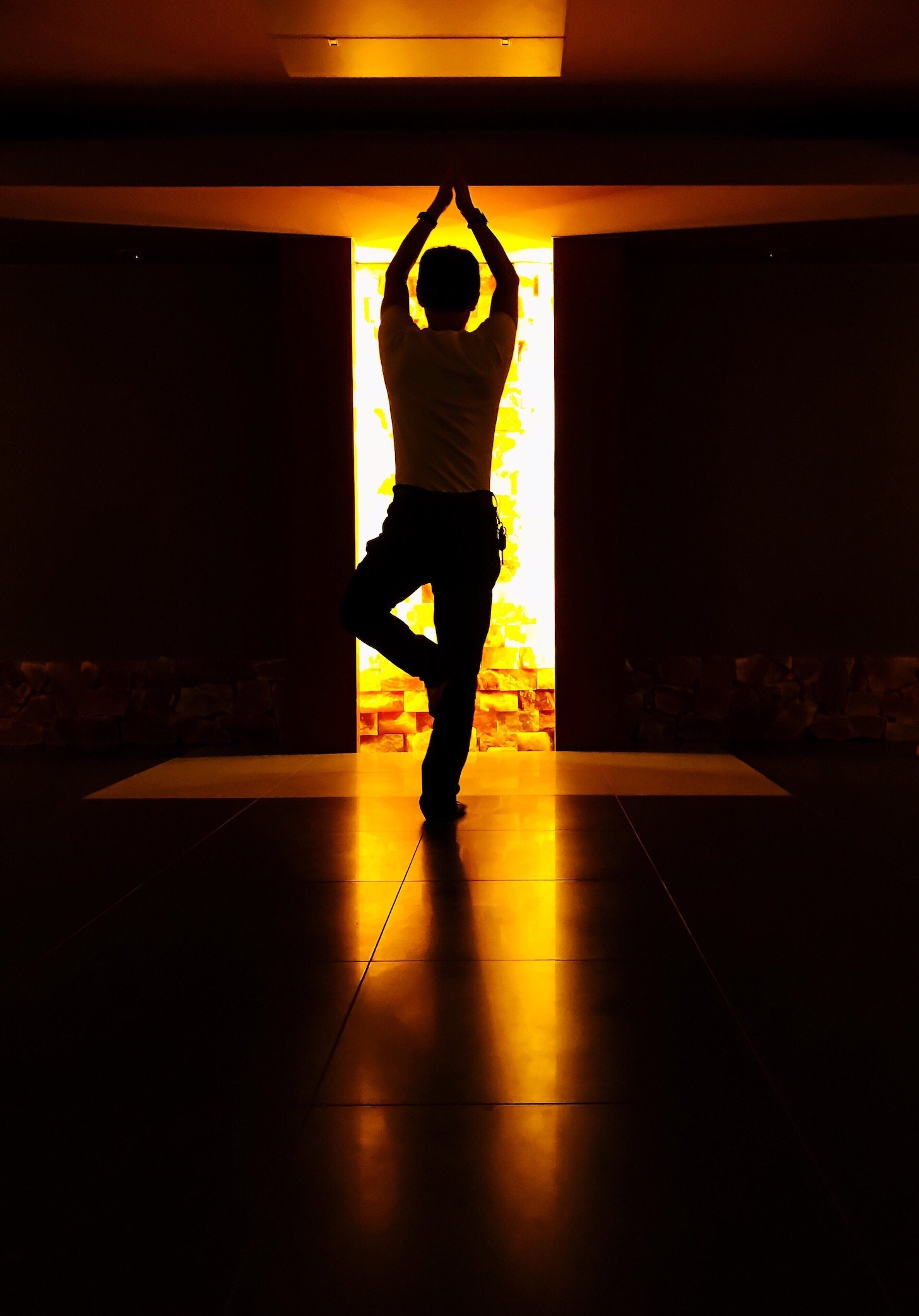 A person doing a yoga tree pose in the dark with a yellow light source entering through a door.