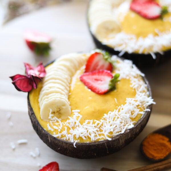 Perspective image of bowls of yellow smoothie bowls topped with coconut shreds, banana and strawberries.