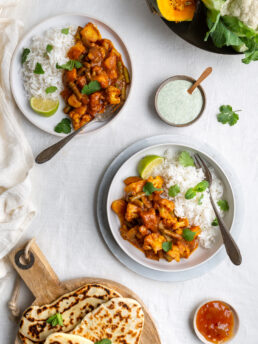 Top down view of a vegetarian, vegan Indian Balti curry with cauliflower and pumpkin served with raita, naan bread and mango chutney and basmati rice on a white background.