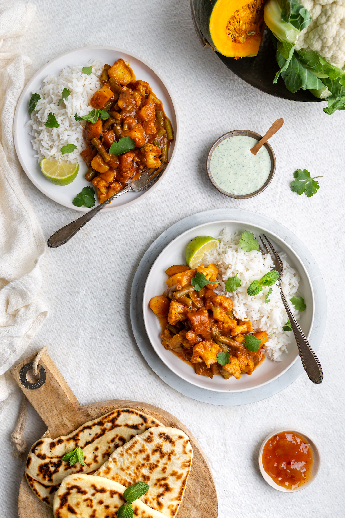 Top down view of a vegetarian, vegan Indian Balti curry with cauliflower and pumpkin served with raita, naan bread and mango chutney and basmati rice on a white background.