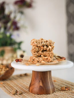 A stack of oat bars topped with peanuts on a cake stand with plants in the background.