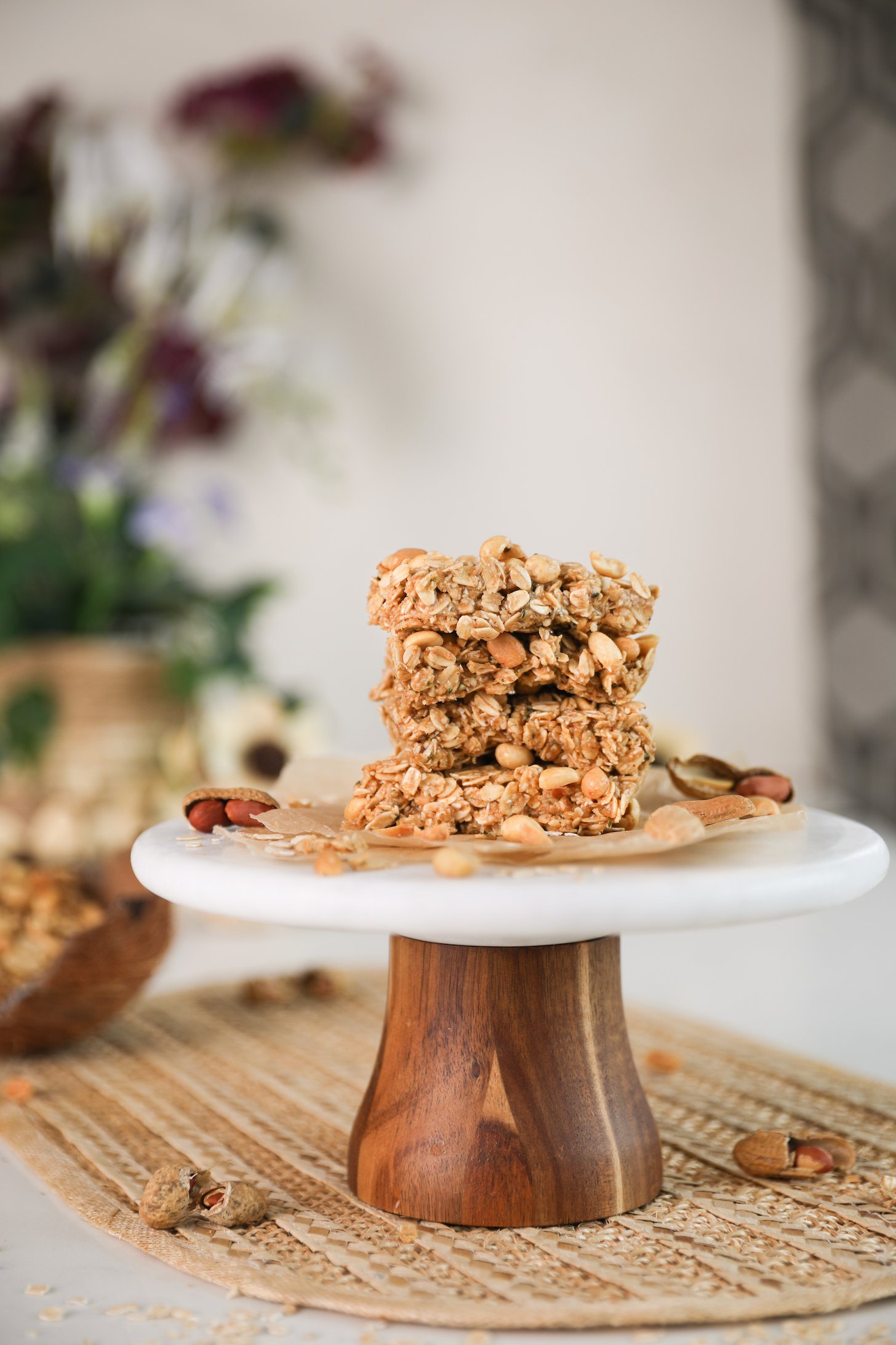 A stack of oat bars topped with peanuts on a cake stand with plants in the background.