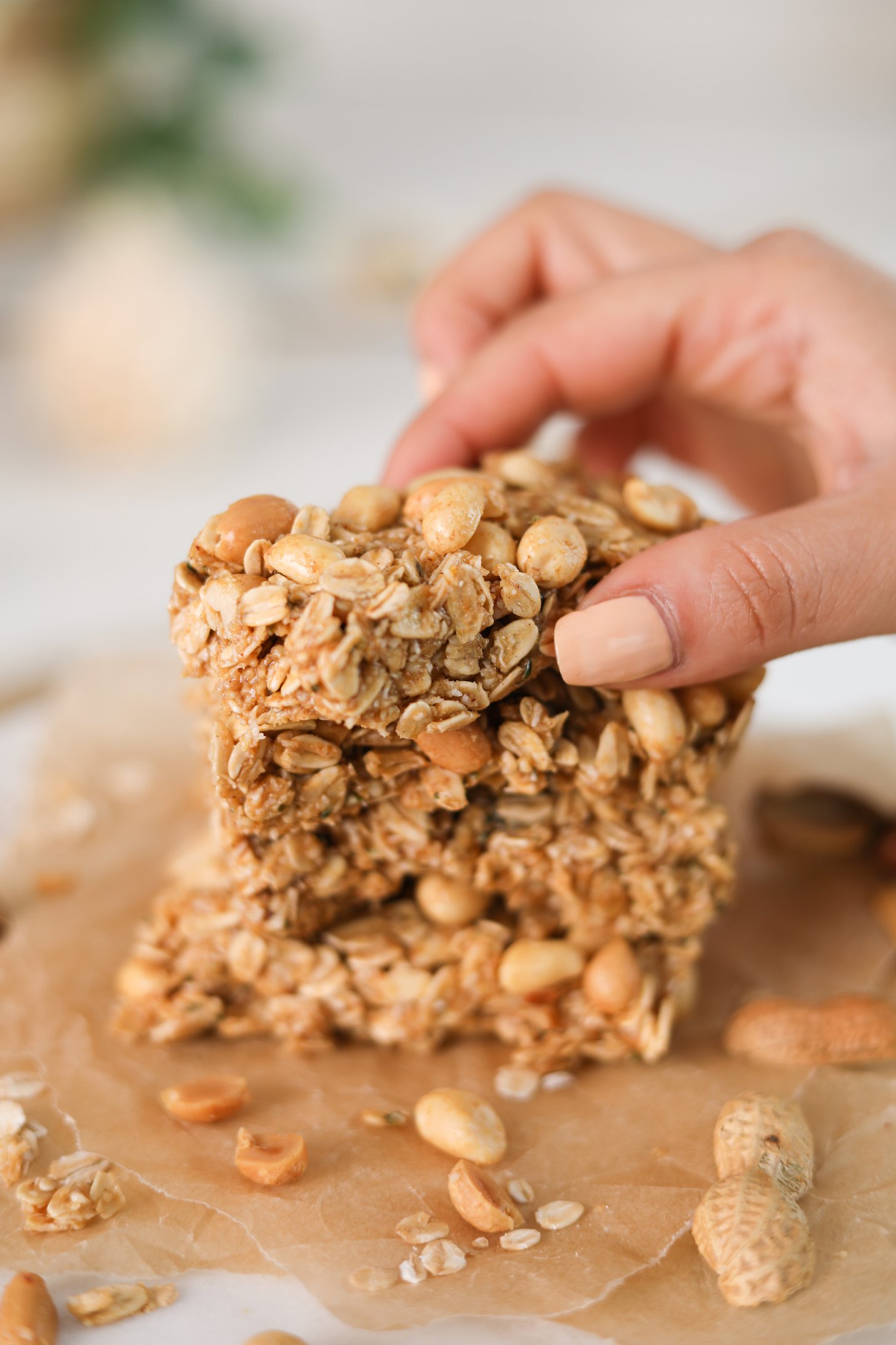 A hand holding an oat bar with peanuts that's on a stack of bars with peanuts all around.