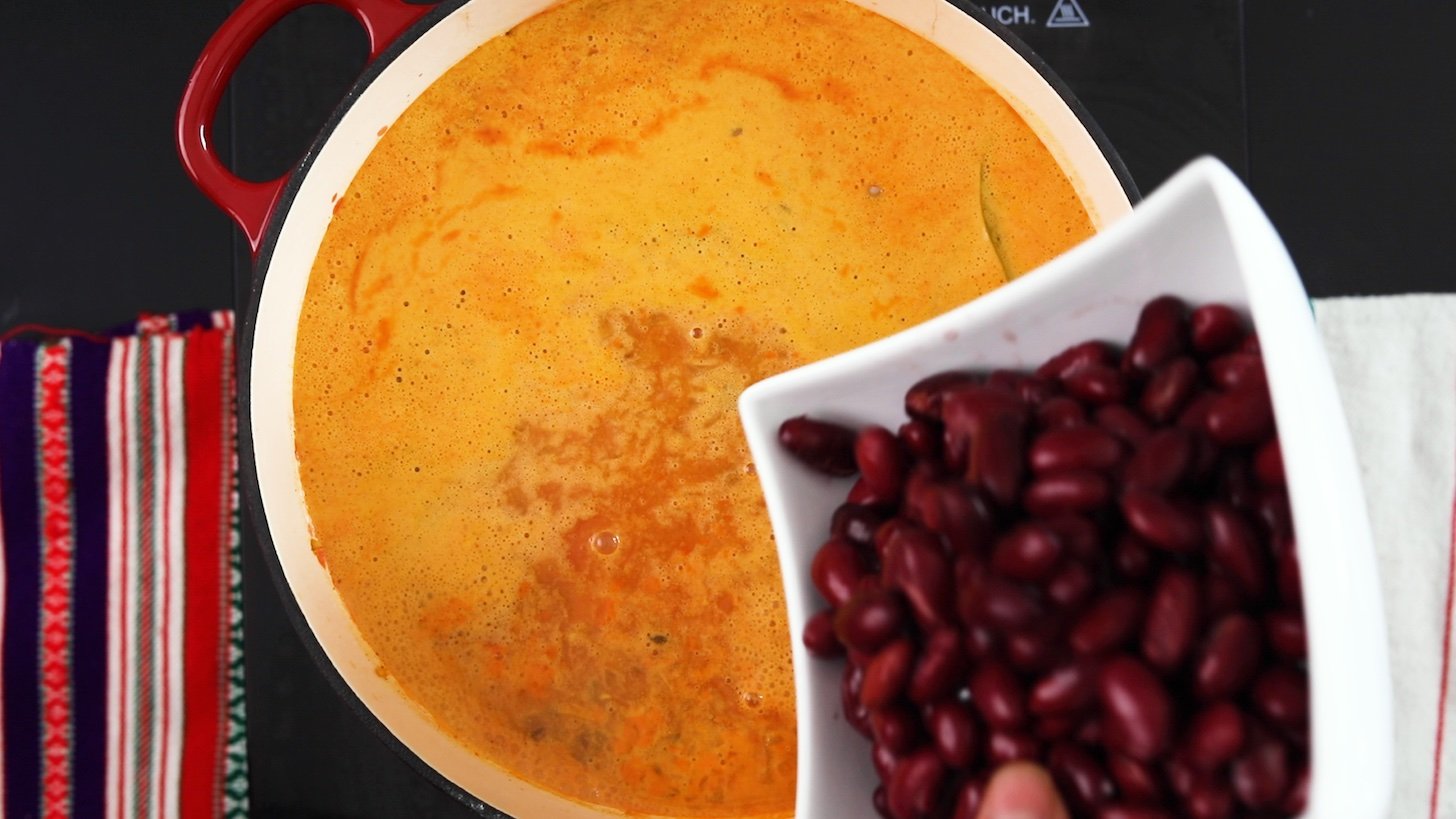 A bowl of kidney beans angled over a cooking pot with an orange liquid in it.