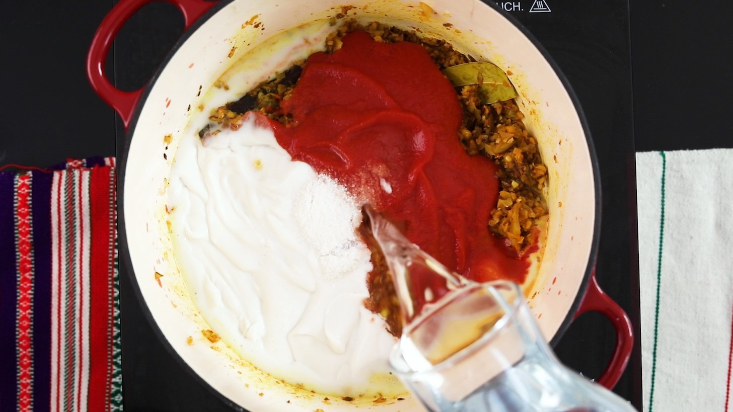 A bottle of water poured into a cooking pot with tomato sauce, coconut cream and spices.