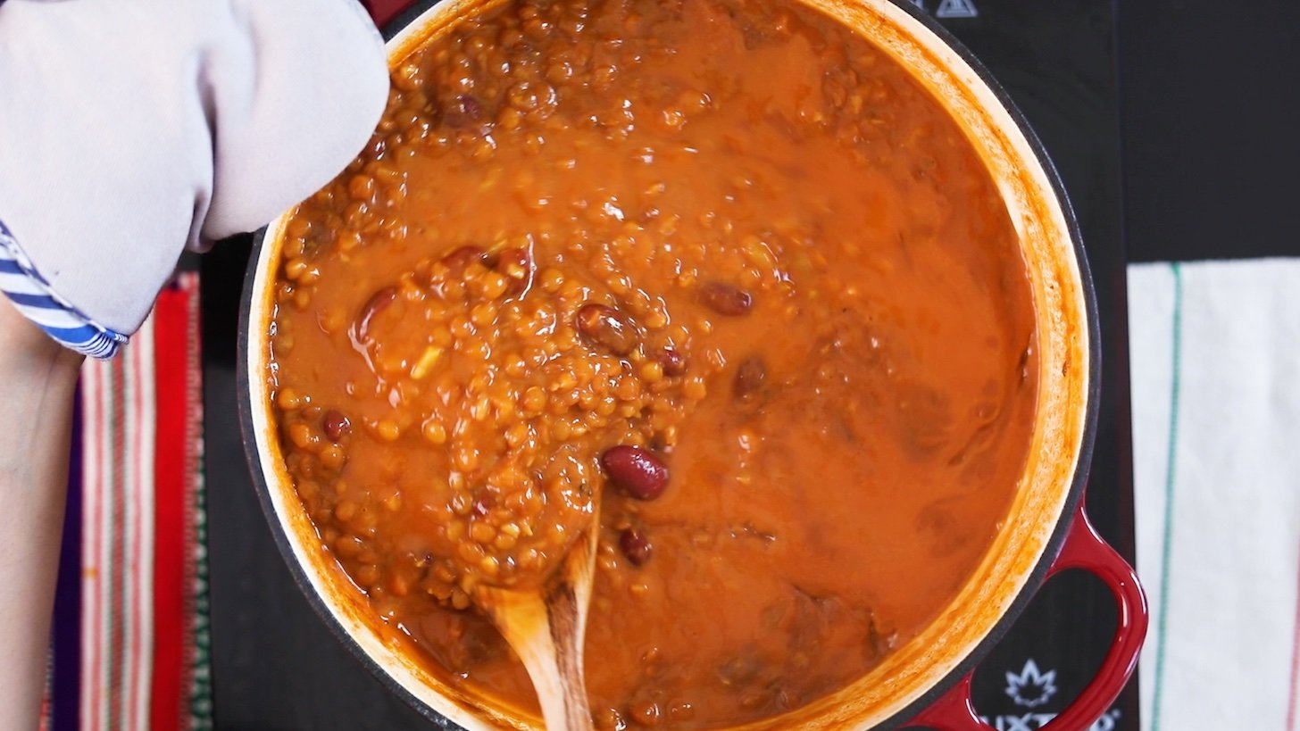 A hand holding a wooden spoon stirring lentil curry in a large cooking pot.