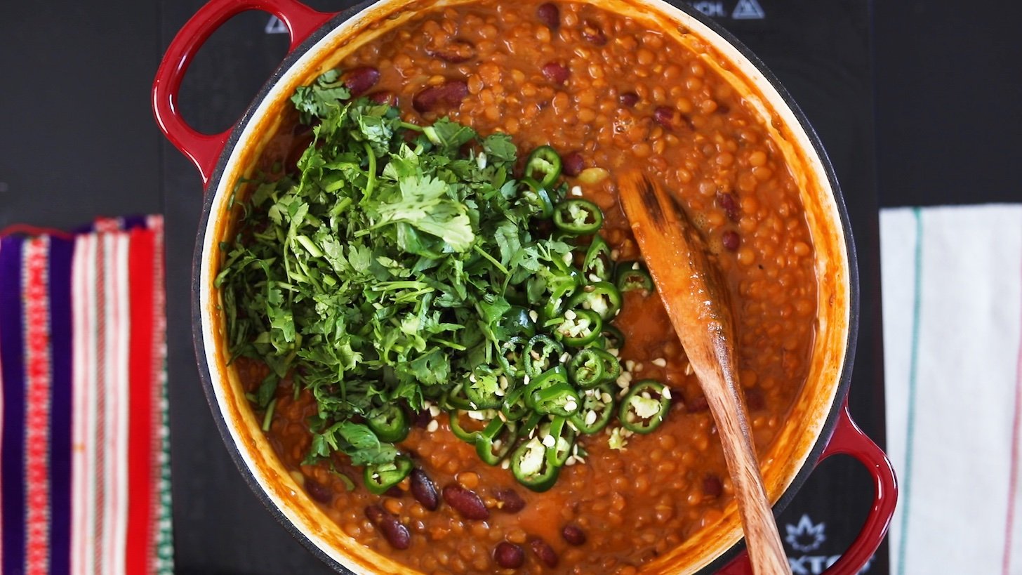 A wooden spoon in a cooking pot filled to the brim with a lentil curry topped with herbs and chillies.