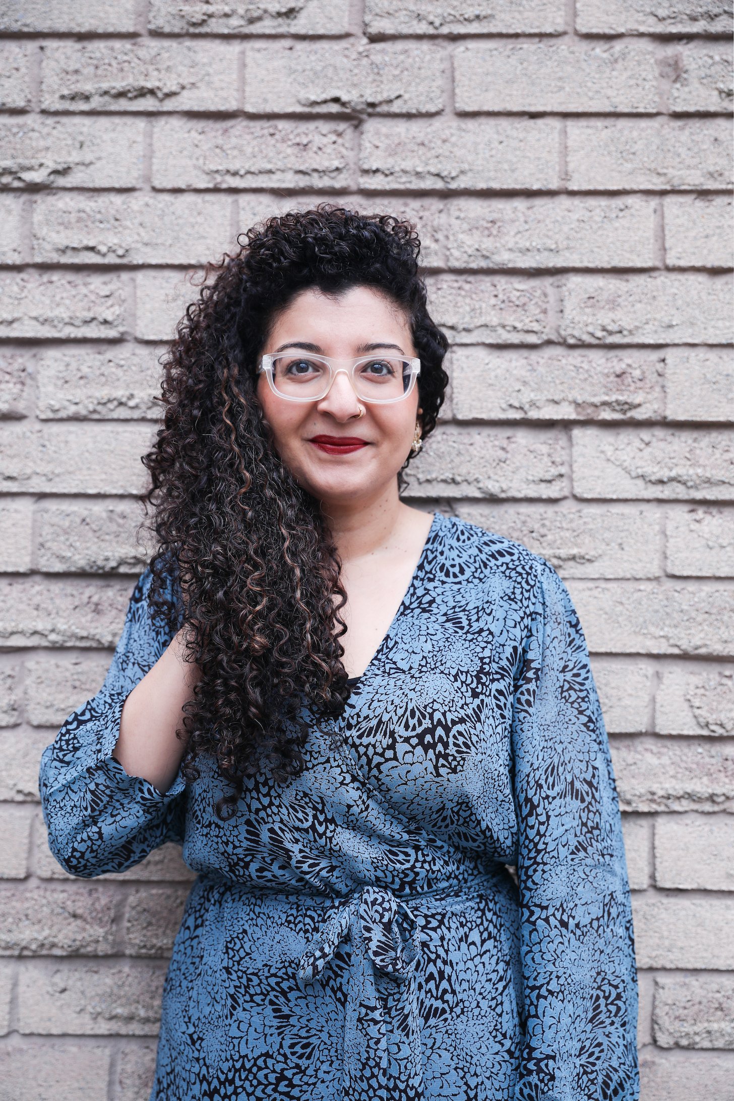 A curly haired woman touching her hair while posing in a printed blue dress against a brick wall.