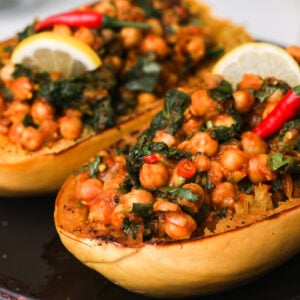 An oven tray with two cooked spaghetti squash halves filled with chickpea and greens curry topped with a lemon slice and red chillies.