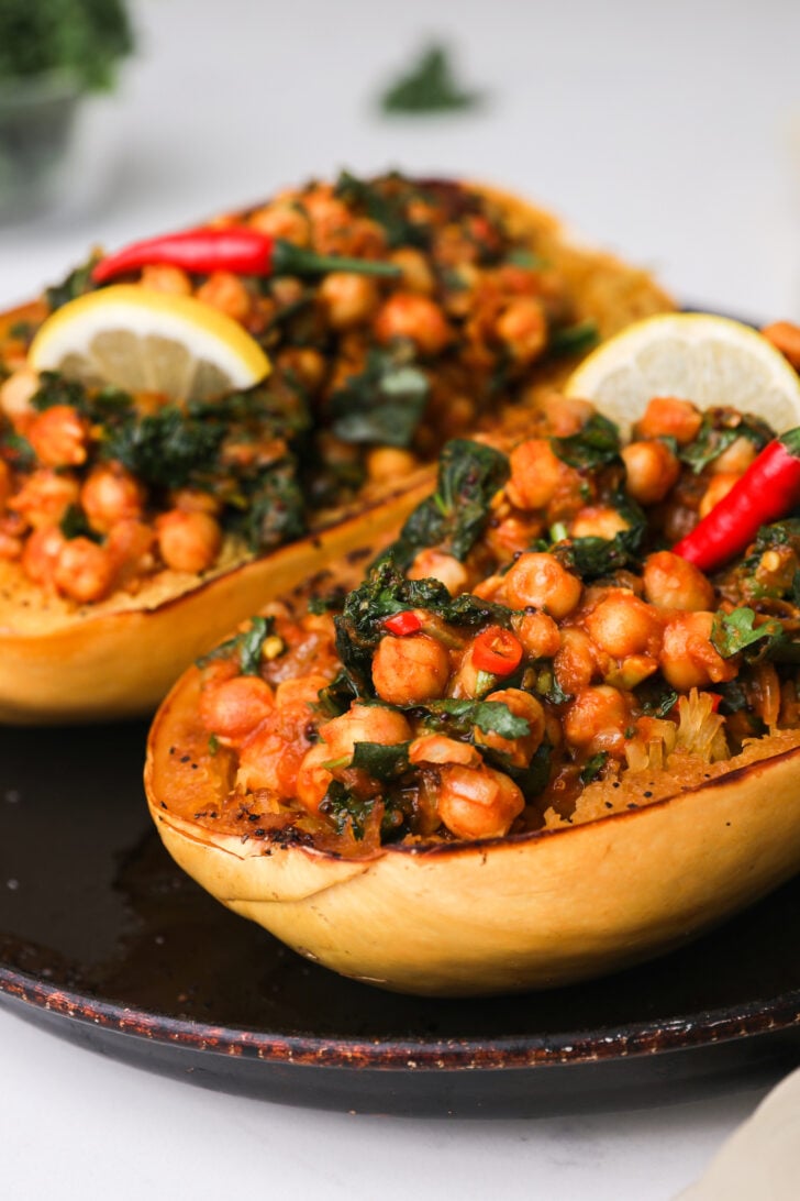 An oven tray with two cooked spaghetti squash halves filled with chickpea and greens curry topped with a lemon slice and red chillies.