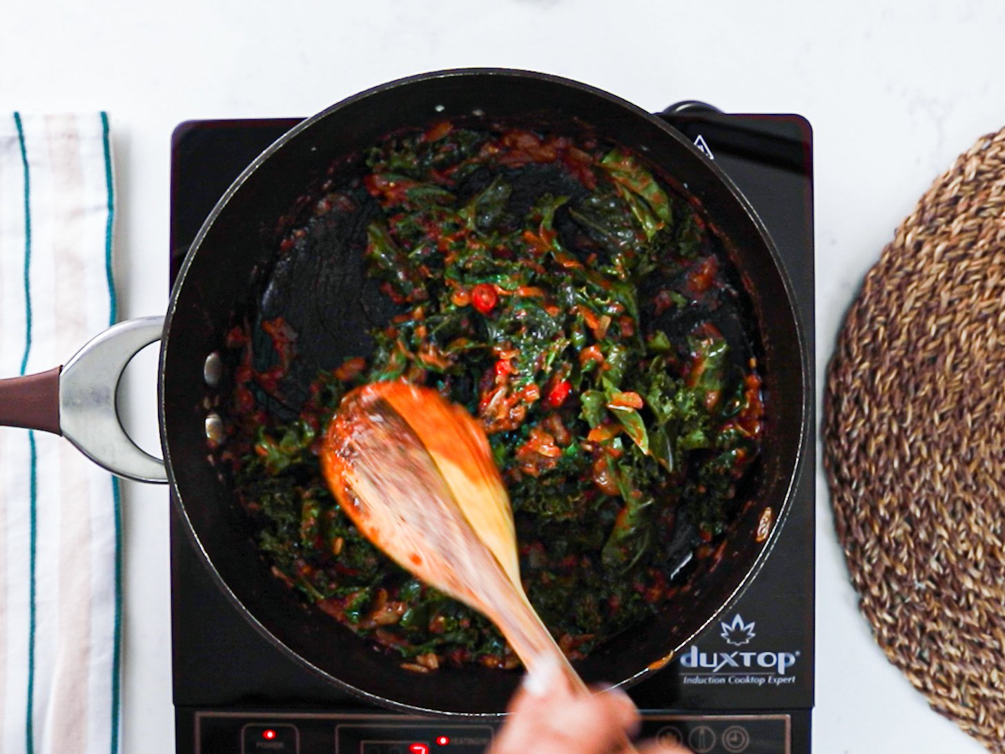 A hand stirring fresh kale in a pan with tomato sauce.