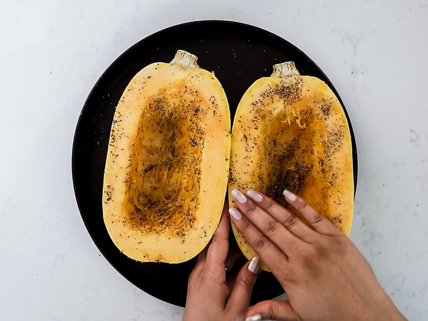 Hands spreading oil, salt and pepper over one half of a spaghetti squash.